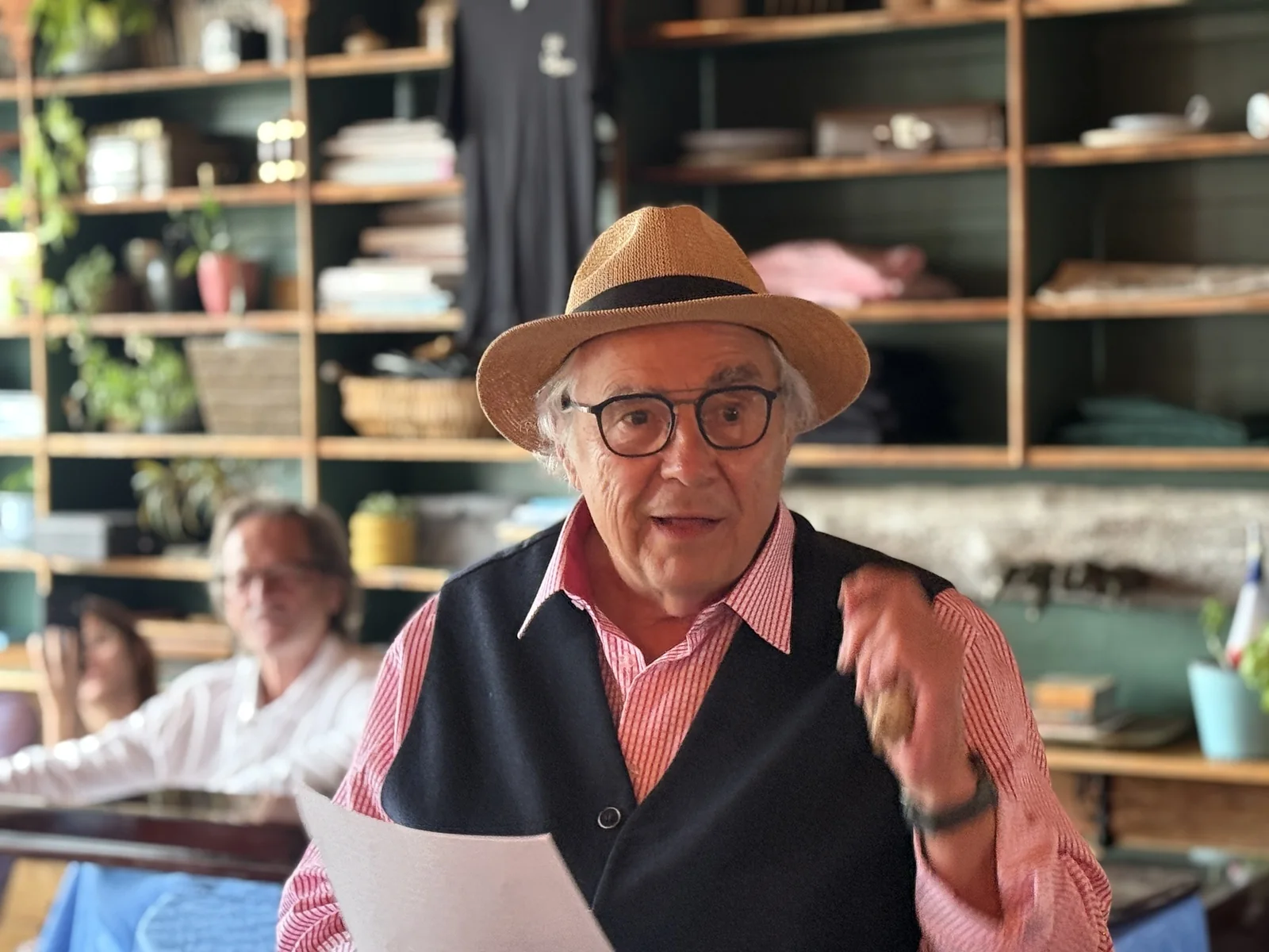 FRED (Denis Fréchette), chapeau de paille et gilet sombre, une feuille à la main, s'adressant à la salle.