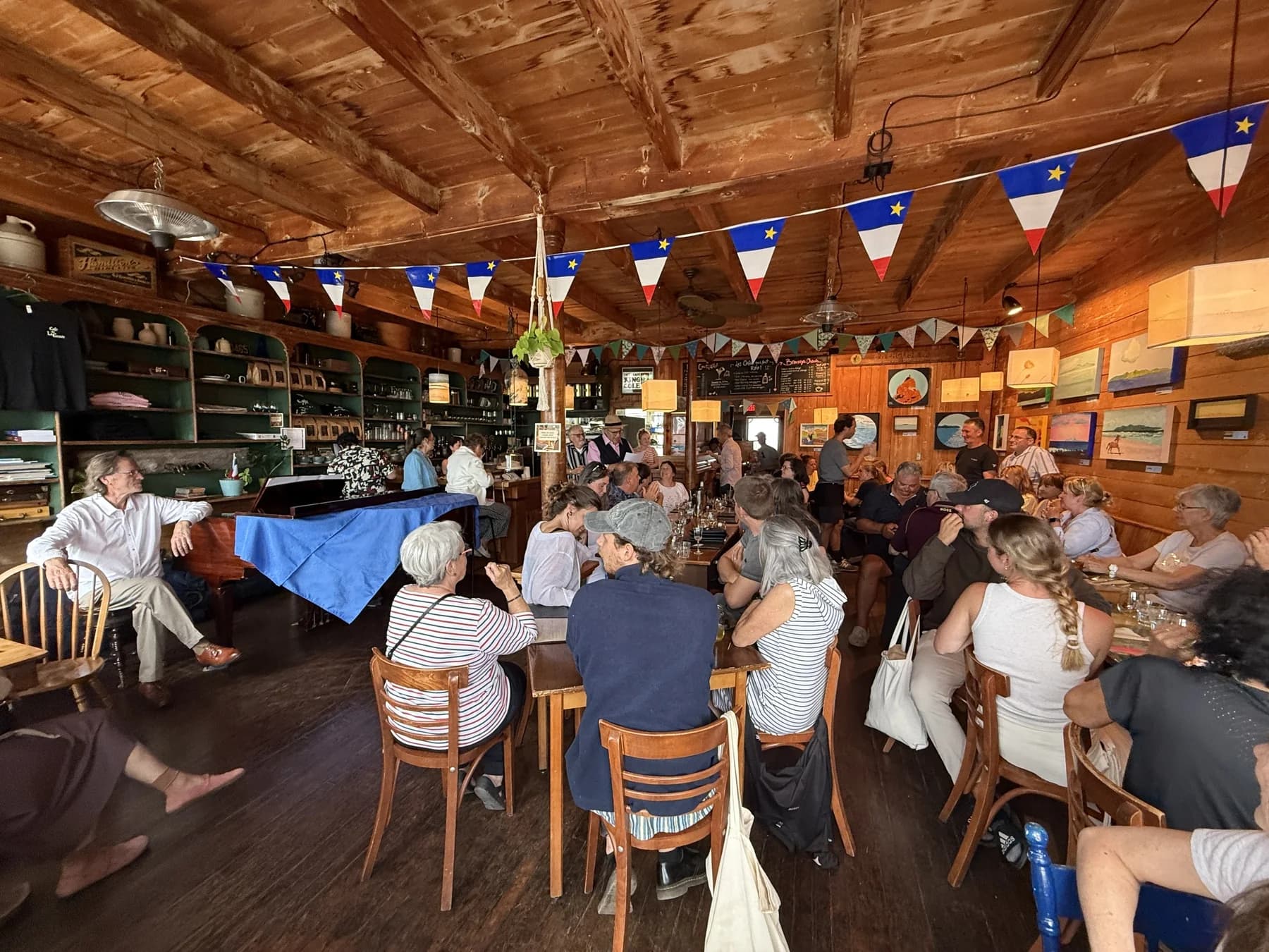La salle du Café de la Grave pendant le vernissage : longues tables occupées, murs de bois clair couverts de toiles, guirlande de fanions acadiens sous la charpente.